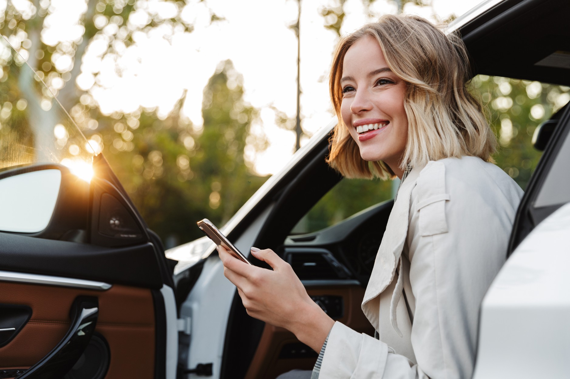 Smiling self-employed woman stepping out of a white car while checking her phone, representing car insurance for freelancers in South Africa